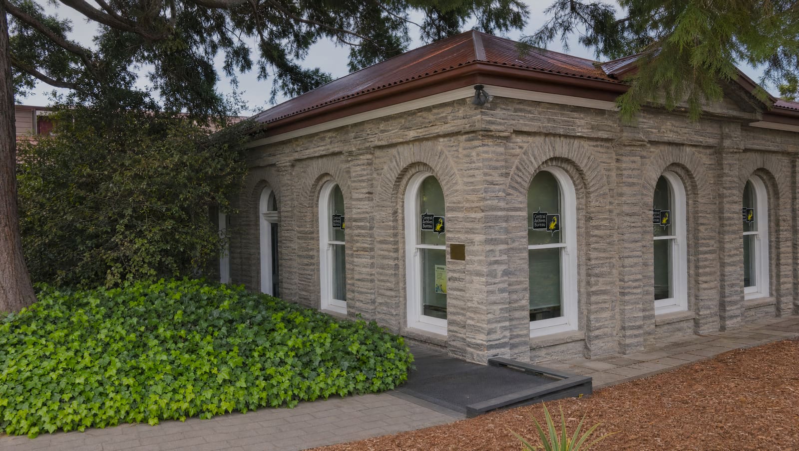 The former Queenstown library of 1877, a single-storey schist building with arched sash windows and a red corrugated iron roof, under a large tree with ivy beside the entrance ramp