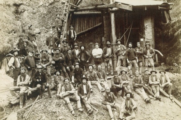 Sepia photograph of about forty bearded miners in hats and waistcoats, standing and sitting on the ground in front of a timber mine entrance and headframe, two dogs among them
