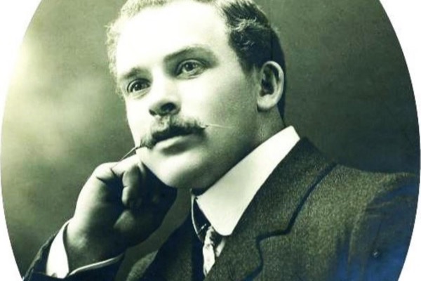 Studio portrait in an oval frame of a young man with fair wavy hair and a moustache, in a dark suit and high collar, his chin resting on his hand