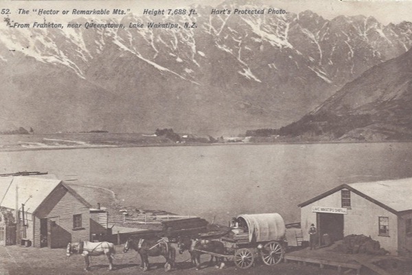 Postcard view across Lake Whakatipu to the snow-streaked Remarkables, with a shed lettered Lake Wakatipu Shipping Co, a second shed, a four-horse team and a canvas-topped wagon drawn up on the shore in the foreground