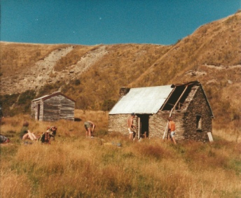 Two stone and timber huts on a tussock hillside, with several people working in the grass outside them.
