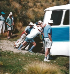 A group of people pushing a blue-and-white campervan stuck on a rough track.
