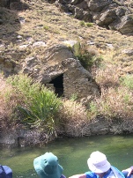 A small arched mine tunnel entrance cut into a rocky riverbank, seen from a boat with passengers' hats in the foreground.