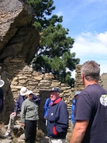 A group of people standing among stone ruins under a large rock overhang, with a lone pine tree above.