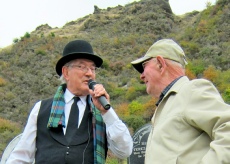 Two men talking, one in a bowler hat and vest holding a microphone, on a hillside among gorse and rock.