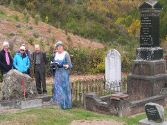 A woman in Victorian dress reading from a paper beside a grave, with a small group looking on among headstones.