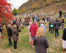 A large crowd gathered among the headstones of a cemetery below a hillside coloured with autumn foliage.