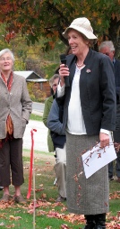 A woman in a cream coat and hat speaking into a microphone in a cemetery.