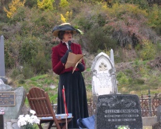 A woman in a dark red jacket and wide-brimmed black hat reading from a folder among gravestones.