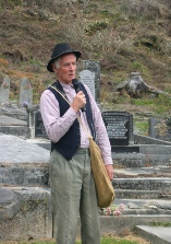 A man in a striped shirt, vest and hat speaking into a microphone, a satchel over his shoulder, beside a gravestone.
