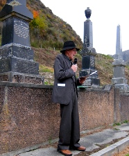 A man in a dark coat and hat speaking beside tall urn-topped gravestone monuments.