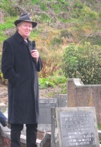 A man in a long dark coat and hat standing beside a fern-covered gravestone.