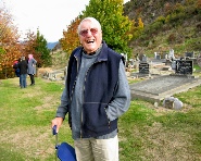 A smiling man in a casual navy jacket holding his hat, with a group of people walking among gravestones behind him.
