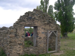 Two people standing inside a roofless stone ruin, framed by its doorway, with willow trees behind.