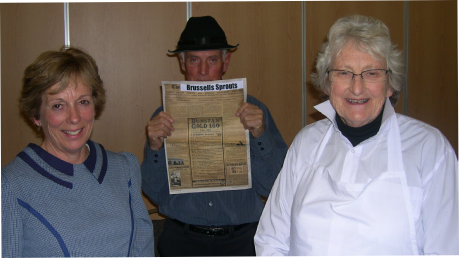 Two women and a man indoors, the man holding up a newspaper page with a large 'Brussells Sprouts' headline.