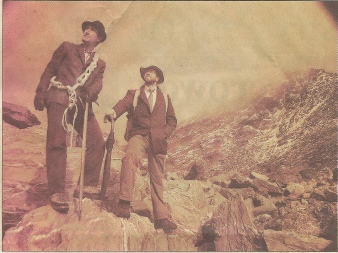 A sepia photograph of two people in old-fashioned dress standing on a rocky mountain slope, one holding a coiled rope and ice axe.