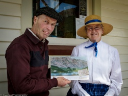 A man and woman in period costume stand on a verandah, the woman holding a framed photograph