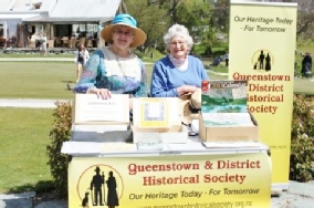 Two women stand behind a trestle table with a Historical Society banner and postcards at an outdoor stall