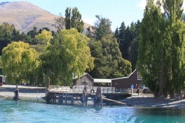 A lakeside jetty and wooden building framed by willow trees on a sunny day.