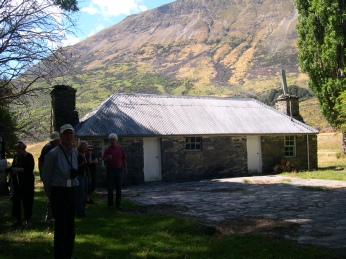 A group of people standing beside an old stone hut with a tussocked mountain behind.