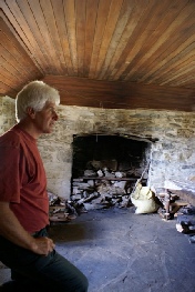 A man standing inside a stone building looking at an old open fireplace.