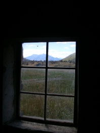 A view through a small stone-walled window over grassland to distant mountains.