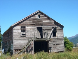 An old weathered wooden barn with a hayloft door, standing in long grass under a blue sky