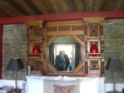 An ornate carved wooden mirror surround built into a stone wall, with two small figures reflected in the mirror and red-shaded lamps on either side