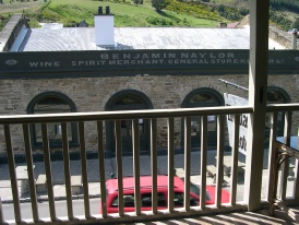 A verandah view of a heritage stone building with painted lettering reading 'Benjamin Naylor, Wine & Spirit Merchant, General Storer', hills rising behind