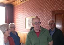 A man in glasses speaking to a small group in a wallpapered room, a framed picture on the wall behind him