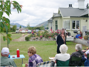 A woman standing and speaking to a seated group on a lawn, with a house and distant mountains behind them