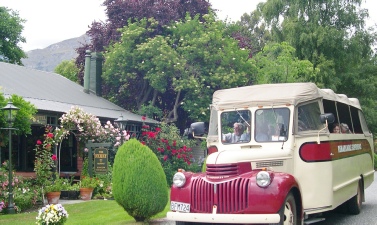 A vintage cream-and-red bus lettered 'Routeburn', parked in a garden beside a cottage with climbing roses