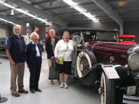 A group of five people stand beside a vintage burgundy touring car inside a museum hall