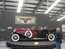 A black and red vintage sports car displayed on a podium inside a museum, with a person looking on