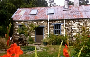 A small stone cottage with a corrugated-iron roof and skylights, orange day lilies growing in front