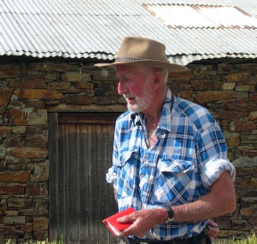 An older man in a wide-brimmed hat and checked shirt standing and talking in front of a stone building with a red door