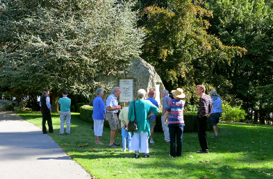A group of people gathered around a memorial plaque mounted on a large rock, under autumn-coloured trees