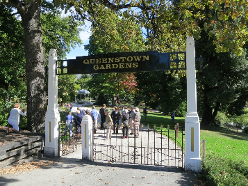 An ornate iron gateway with an overhead sign reading 'Queenstown Gardens', people walking through beneath autumn trees