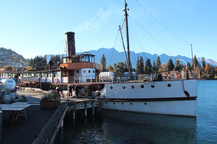The TSS Earnslaw alongside the Queenstown wharf on a clear day, her red funnel and white hull against the Remarkables, members boarding