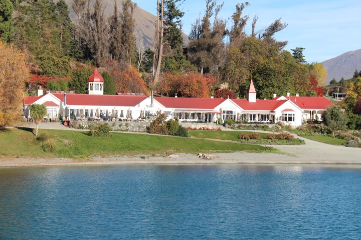 The white Walter Peak homestead with its red roofs and turrets seen across the lake, autumn trees behind and a lawn down to the beach