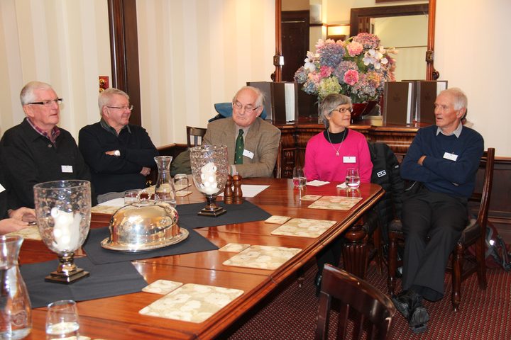 Five members seated along one side of a long polished table set with silver cloches and place mats, a vase of hydrangeas behind them