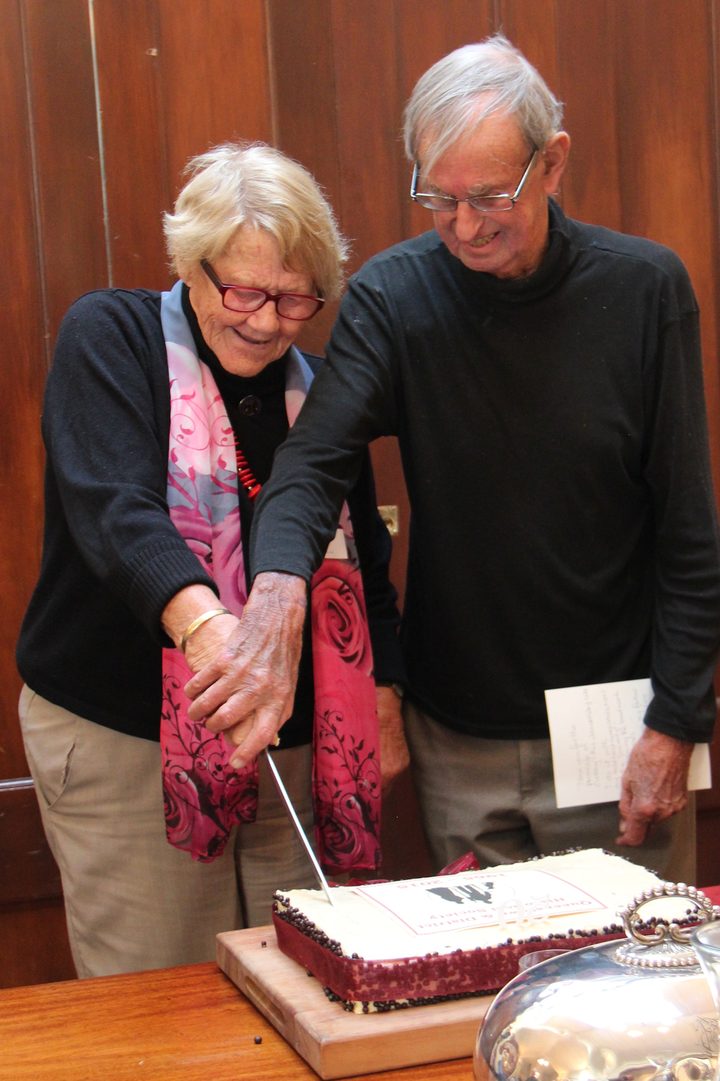 A woman in a pink floral scarf and a man in glasses cutting the Society’s anniversary cake together, both hands on the knife