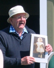 An elderly man in a white sun hat holding up an old family photograph while speaking outdoors.
