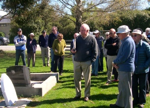 A group of about twenty people standing among headstones in a cemetery, listening to a man giving a talk.