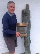 A man in a blue jumper holding an old wooden and metal cask beside a weathered post against a white wall