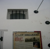 Exterior wall of the Arrowtown Gaol with a small barred window and a 'The Law Breakers' exhibition poster mounted beside the door