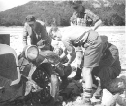 Vintage black-and-white film still of several men and boys working on a vintage car's engine beside a riverbed