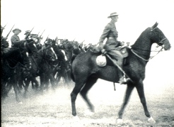 Black-and-white archival film still of a mounted officer on horseback leading marching soldiers