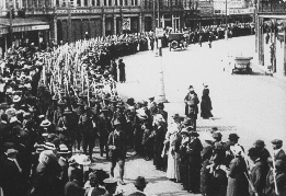 Black-and-white archival photograph of soldiers marching through a town street past crowds of onlookers