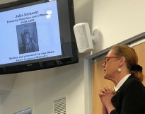 A woman stands with hands clasped beside a screen showing a portrait of Julia Eichardt, written and presented by Ann Mann